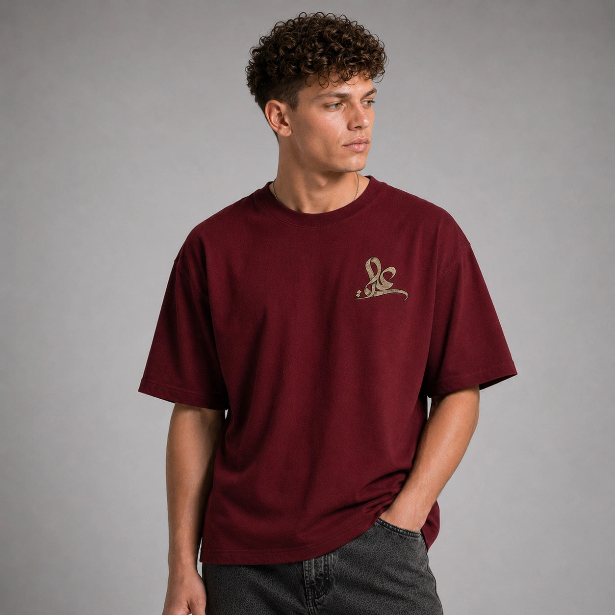 A man wearing a maroon short-sleeve t-shirt with a small gold embroidered logo on the left chest, paired with dark gray jeans, photographed against a neutral gray studio background.