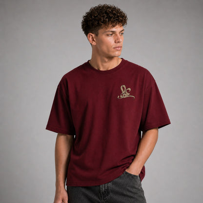 A man wearing a maroon short-sleeve t-shirt with a small gold embroidered logo on the left chest, paired with dark gray jeans, photographed against a neutral gray studio background.