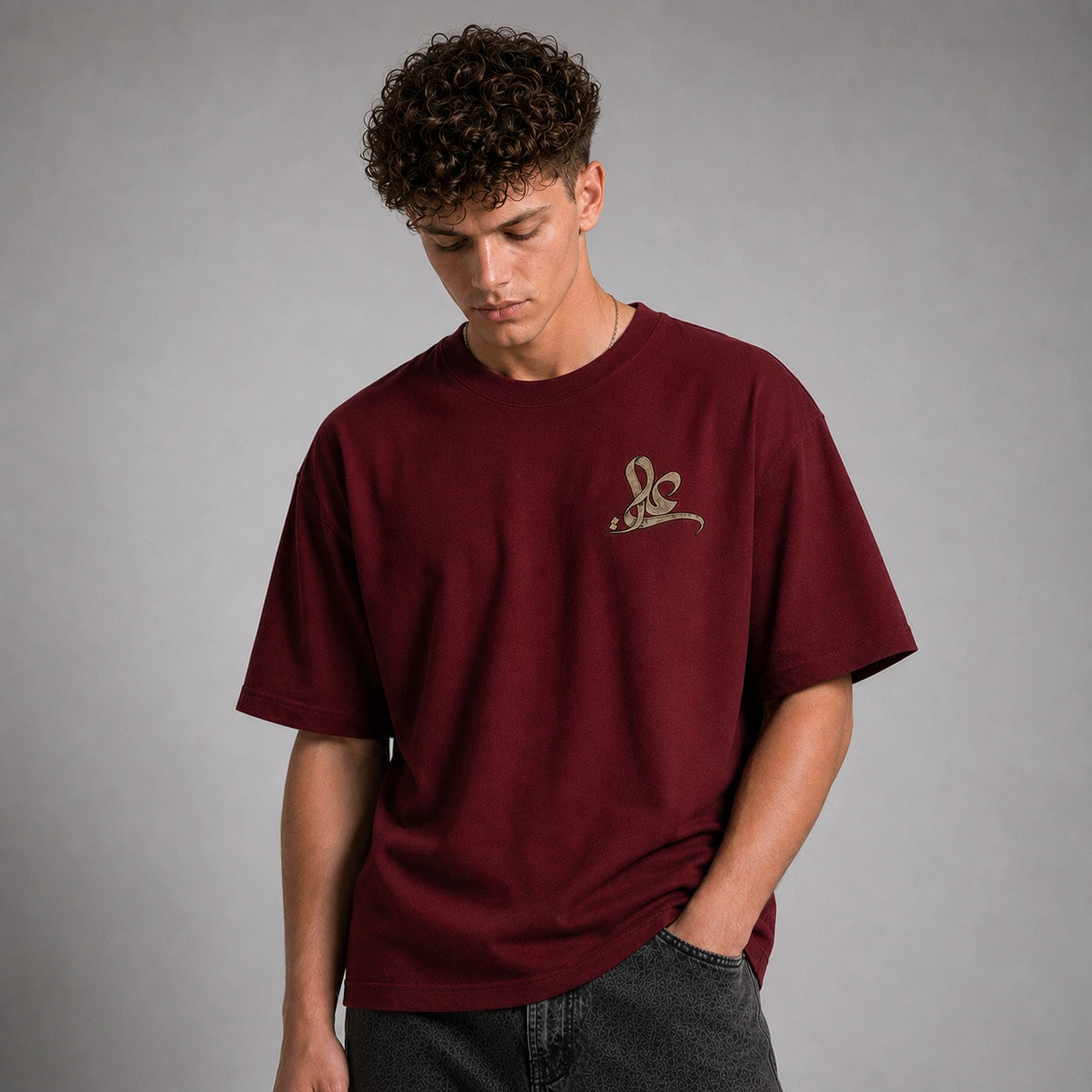 A man wearing an oversized maroon t-shirt with a small gold embroidered logo on the left chest, paired with dark gray jeans, set against a plain gray studio background.