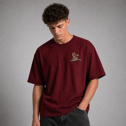 A man wearing an oversized maroon t-shirt with a small gold embroidered logo on the left chest, paired with dark gray jeans, set against a plain gray studio background.