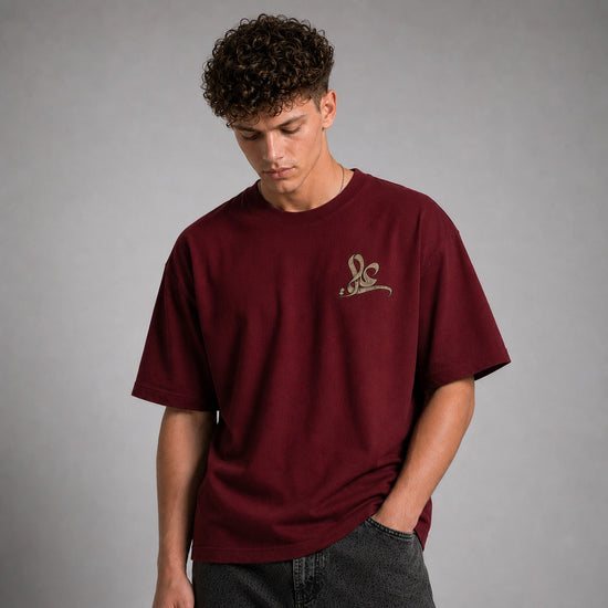 A man wearing an oversized maroon t-shirt with a small gold embroidered logo on the left chest, paired with dark gray jeans, set against a plain gray studio background.