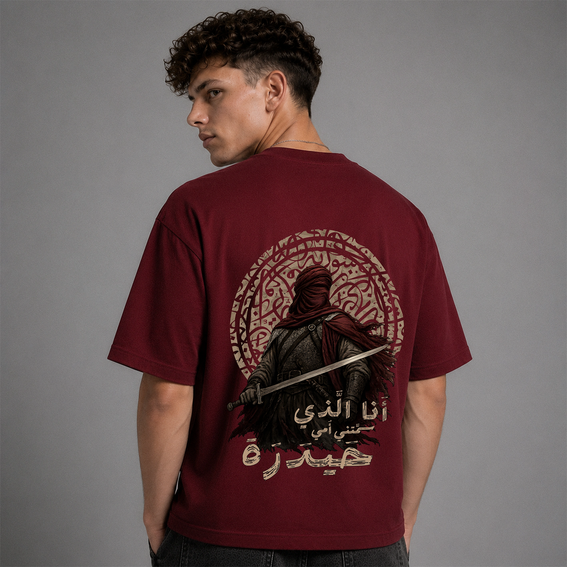 A man shown from the back wearing a maroon short-sleeve graphic T-shirt with a large printed design featuring a hooded figure, Arabic text, and circular decorative elements on a plain gray studio background.