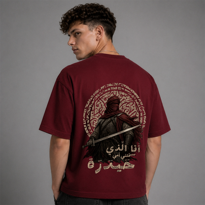 A man shown from the back wearing a maroon short-sleeve graphic T-shirt with a large printed design featuring a hooded figure, Arabic text, and circular decorative elements on a plain gray studio background.