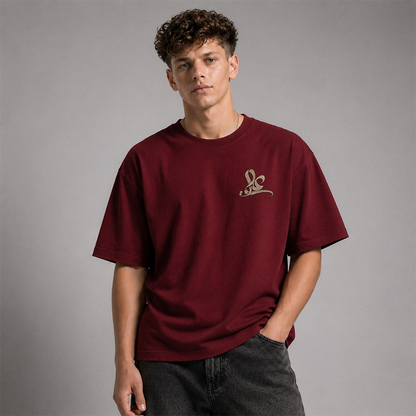 A young man with curly brown hair wearing an oversized maroon crewneck t-shirt with a small gold embroidered logo on the left chest, paired with dark gray jeans, against a solid gray studio background.