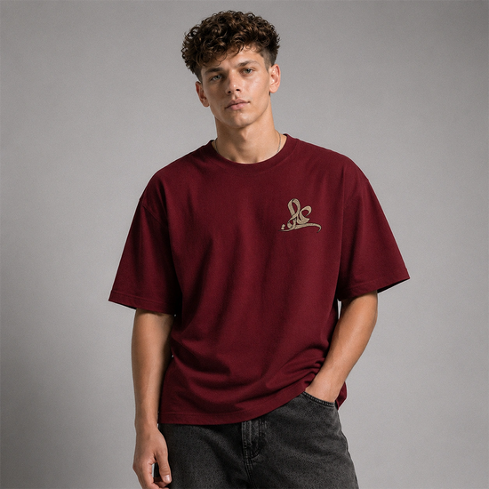 A young man with curly brown hair wearing an oversized maroon crewneck t-shirt with a small gold embroidered logo on the left chest, paired with dark gray jeans, against a solid gray studio background.