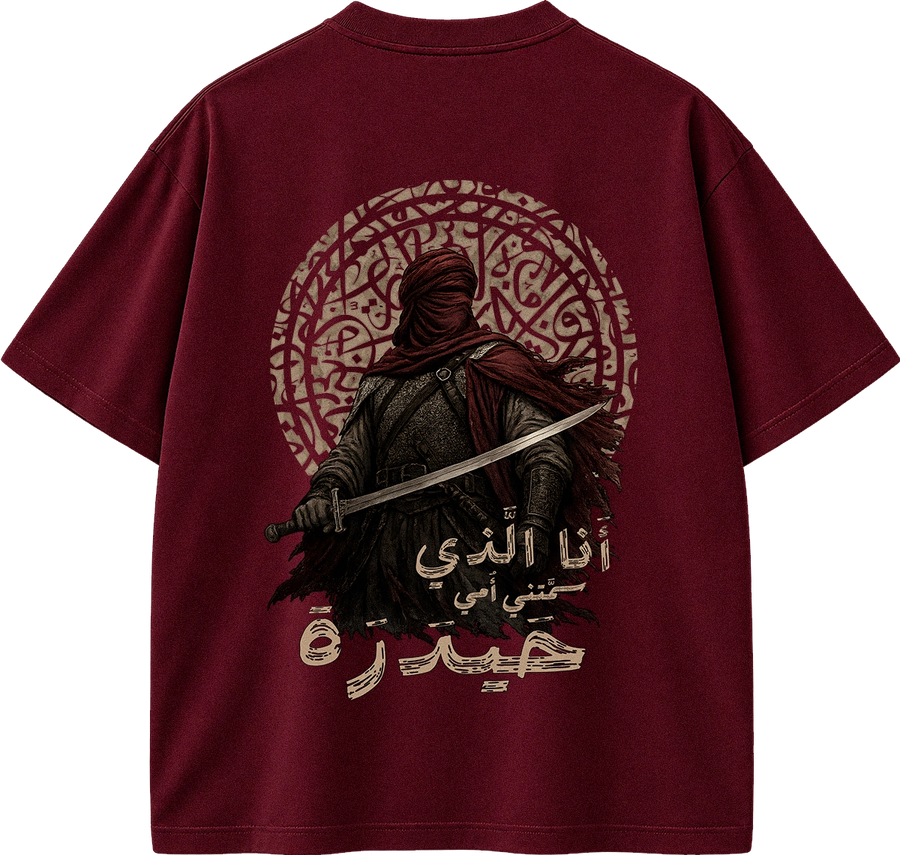 A maroon short-sleeve t-shirt shown from the back, featuring a large graphic print of a hooded figure holding a sword in front of an intricate circular pattern, with Arabic text printed below.