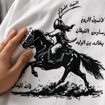 White t-shirt with black graphic of a rider on a horse and Arabic text, held by a hand.