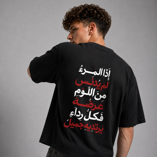 A man wearing a black t-shirt, viewed from behind, showing a large Arabic text graphic printed in white and red on the back, set against a plain gray studio background.