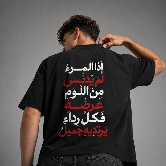 A man viewed from behind wearing a black t-shirt with a large Arabic text graphic printed on the back in white and red, set against a plain gray studio background.