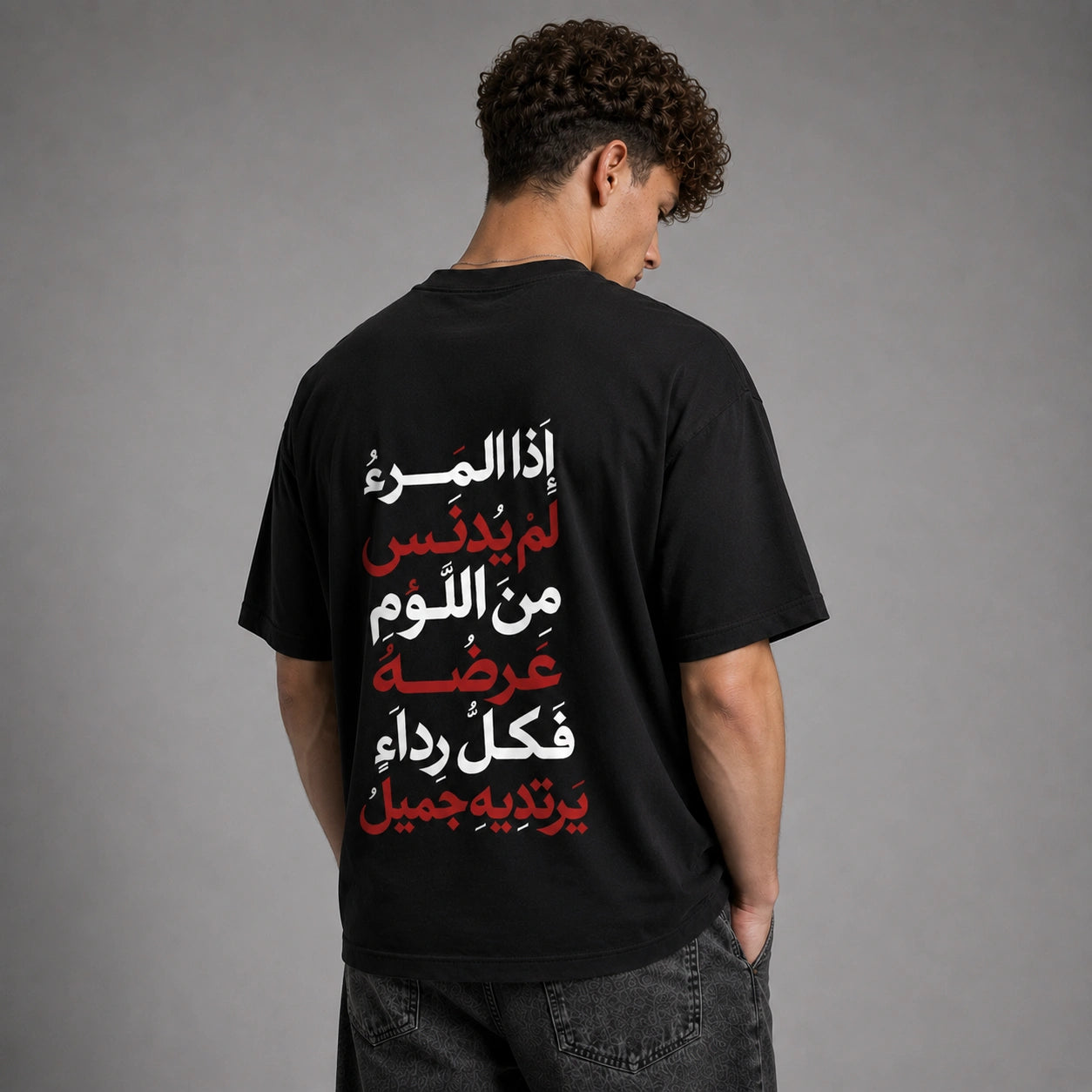 A man viewed from behind wearing a black t-shirt with vertical Arabic text printed on the back in white and red, paired with dark gray jeans against a neutral gray studio background.