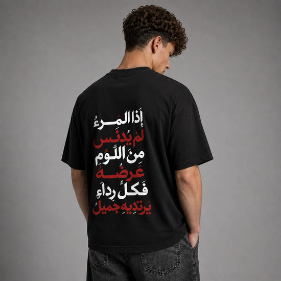 A man viewed from behind wearing a black t-shirt with vertical Arabic text printed on the back in white and red, paired with dark gray jeans against a neutral gray studio background.