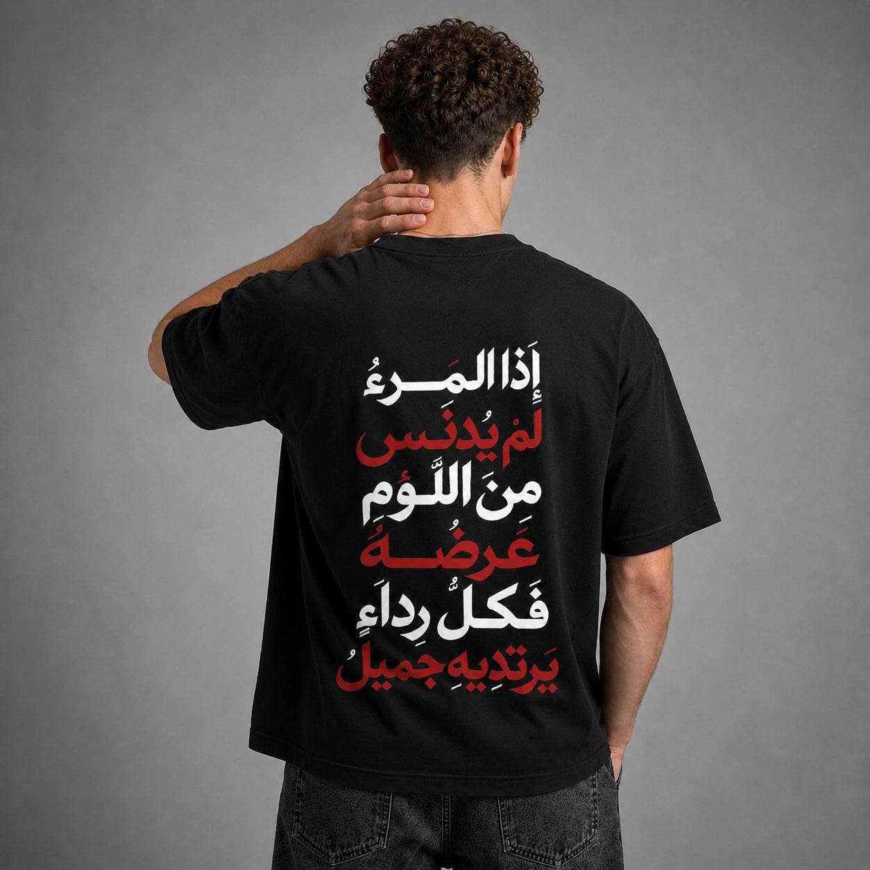 A man viewed from behind wearing a black t-shirt with a large Arabic text graphic printed on the back in white and red, set against a plain gray studio background.