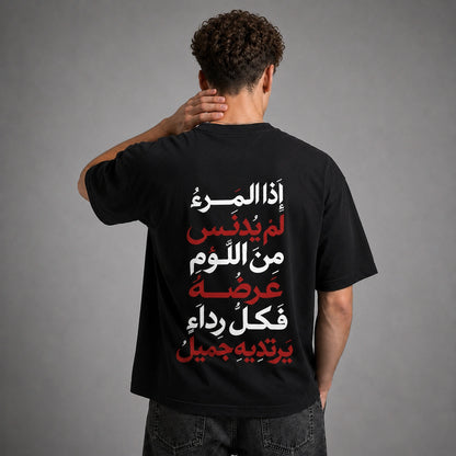 A man viewed from behind wearing a black t-shirt with a large Arabic text graphic printed on the back in white and red, set against a plain gray studio background.