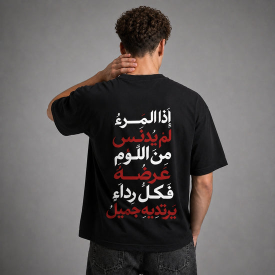 A man viewed from behind wearing a black t-shirt with a large Arabic text graphic printed on the back in white and red, set against a plain gray studio background.