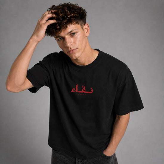 A young man with curly brown hair wearing a black crewneck t-shirt with red Arabic calligraphy on the chest, posing against a solid gray studio background.