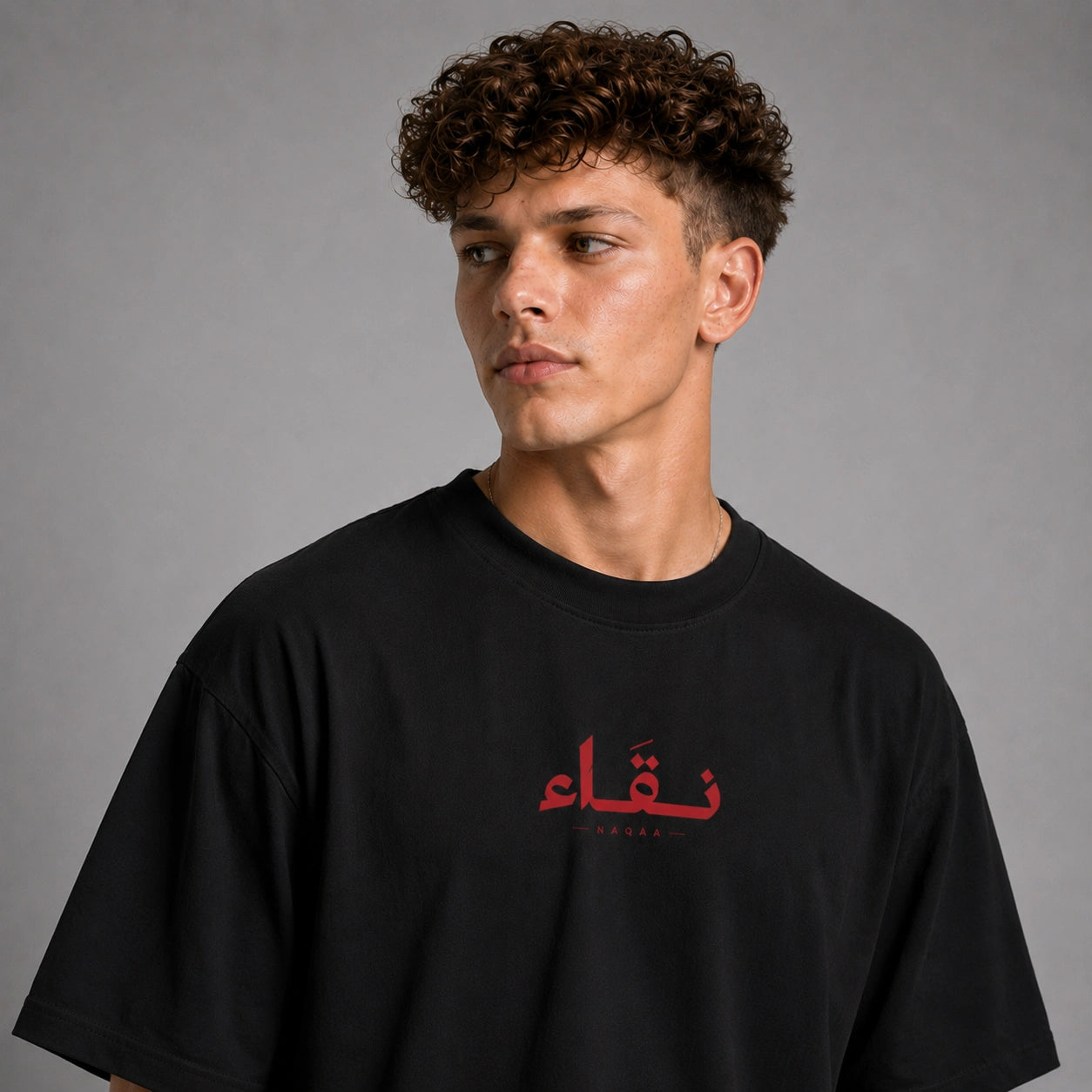 A man with curly brown hair wearing a black oversized t-shirt with red Arabic calligraphy and the word 'SUDAR' printed on the chest, photographed against a neutral gray background.