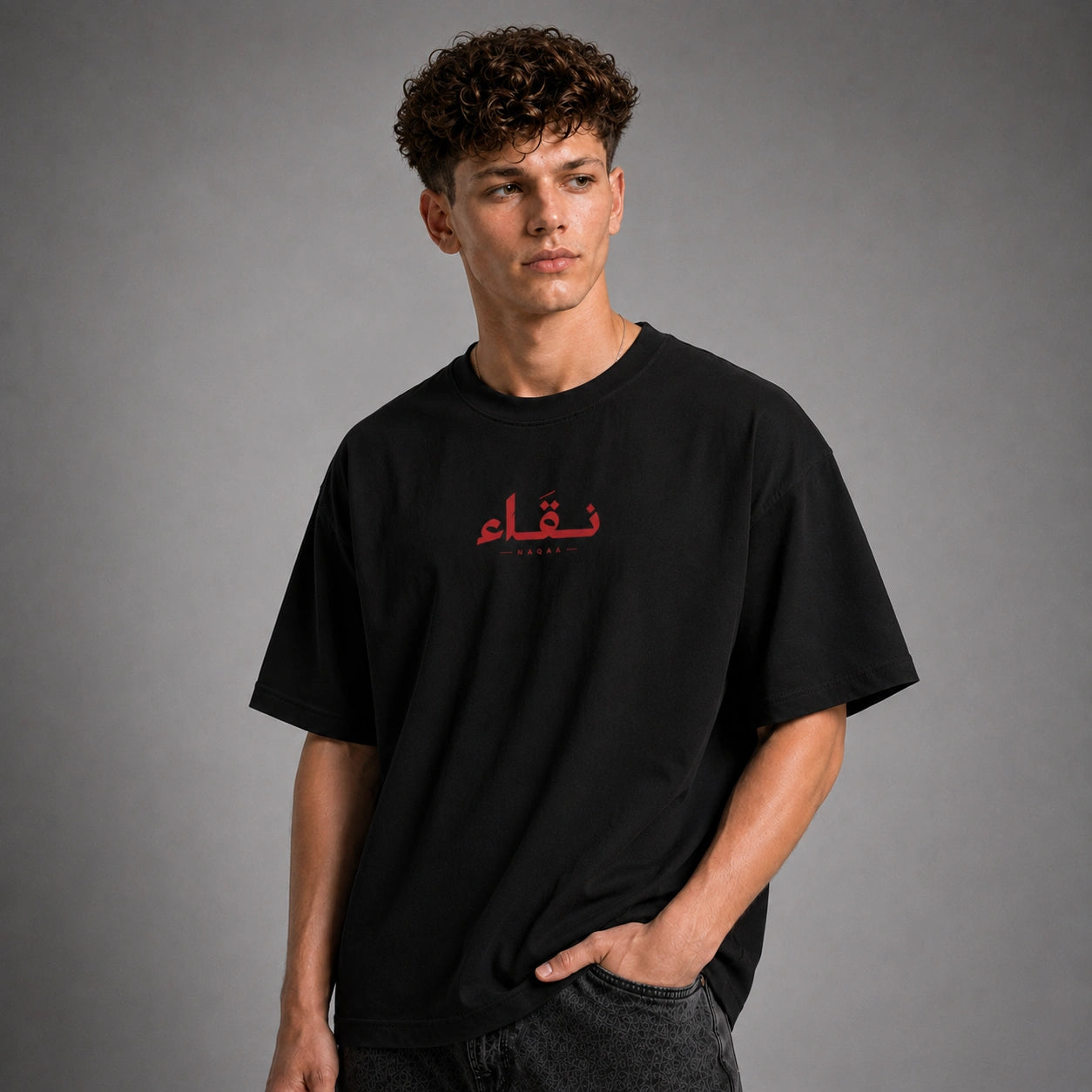 A man wearing a black oversized t-shirt with red Arabic calligraphy on the chest, posed against a neutral gray studio background.