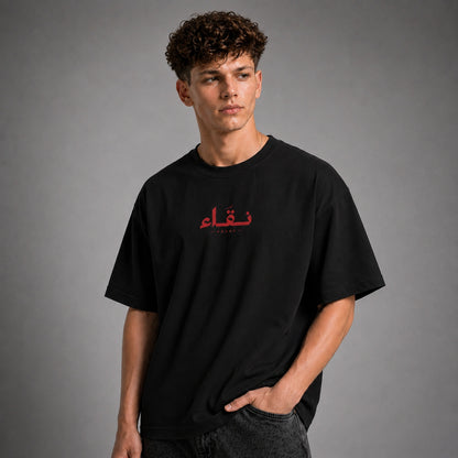 A man wearing a black oversized t-shirt with red Arabic calligraphy on the chest, posed against a neutral gray studio background.