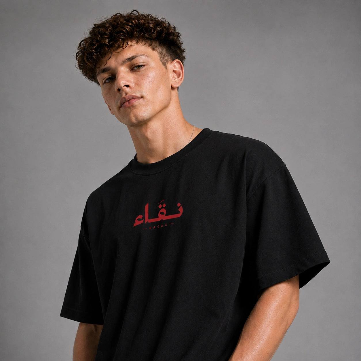 A man wearing a black oversized t-shirt with red Arabic calligraphy on the chest, photographed against a neutral gray studio background.