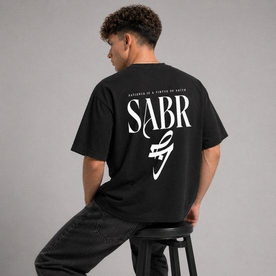 Person wearing a black t-shirt with 'SABR' logo and text on the back, sitting on a stool against a gray background.