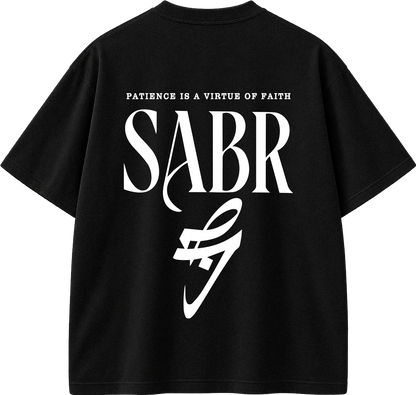 Black short-sleeve t-shirt with a large white graphic print on the back. The design features the text 'PATIENCE IS A VIRTUE OF FAITH' above the word 'SABR' in a stylized serif font, with a white abstract logo underneath.