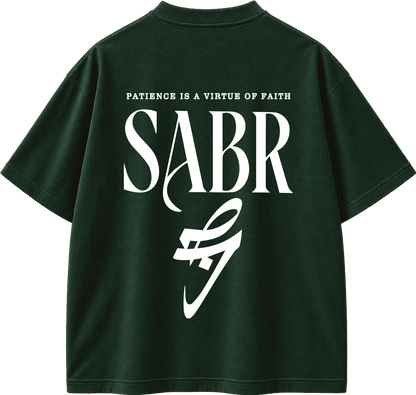 A dark forest green short-sleeve t-shirt shown from the back, featuring a large white graphic print with the text 'PATIENCE IS A VIRTUE OF FAITH' and 'SABR' in a stylized serif font, with a white abstract swoosh-like graphic below.