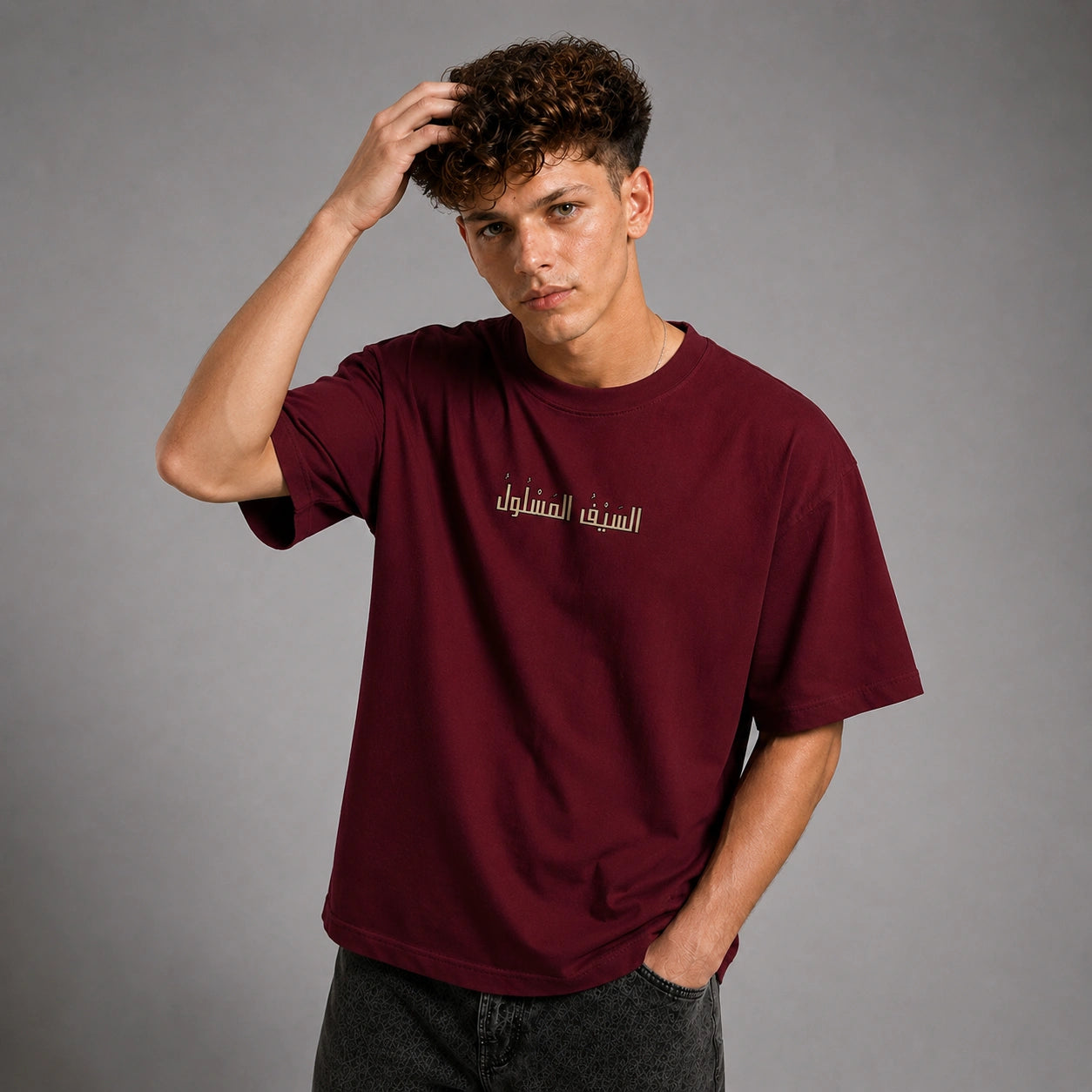 A young man with curly brown hair wearing an oversized maroon t-shirt with white Arabic text on the chest, posing against a solid gray studio background.