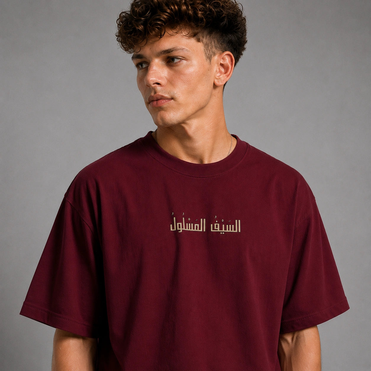 A young man with curly brown hair wearing a maroon crewneck t-shirt with white Arabic text printed on the chest, photographed against a neutral gray background.