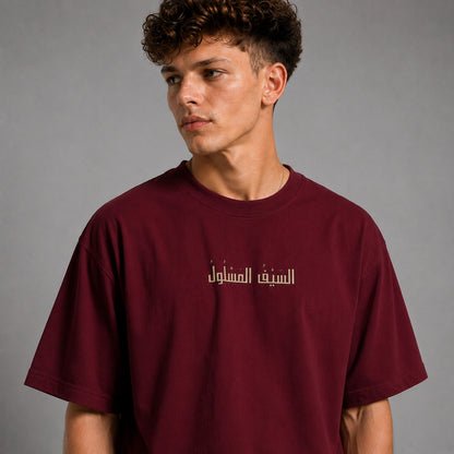 A young man with curly brown hair wearing a maroon crewneck t-shirt with white Arabic text printed on the chest, photographed against a neutral gray background.