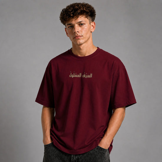 A young man wearing a maroon short-sleeve t-shirt with white Arabic text on the chest, posed against a solid gray studio background.