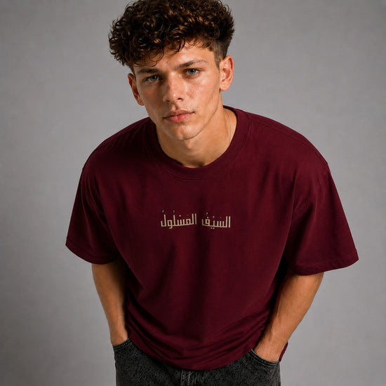 A young man with curly brown hair wearing a maroon crewneck t-shirt with gold Arabic text on the chest, posed against a neutral gray background.