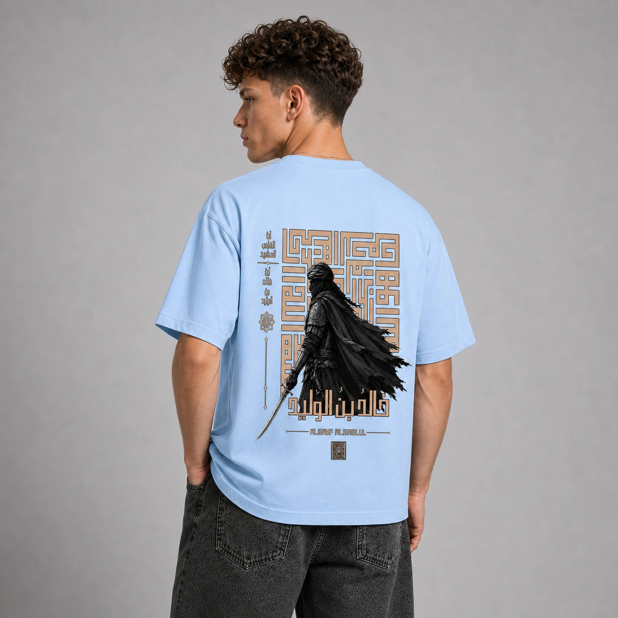 A man wearing a light blue t-shirt from the back, featuring a large graphic print of Darth Vader in black with gold geometric details and text. He is paired with dark gray jeans against a neutral gray studio background.