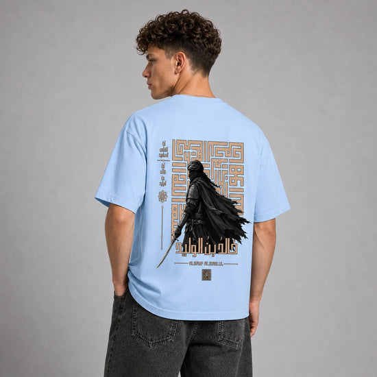 A man wearing a light blue t-shirt from the back, featuring a large graphic print of Darth Vader in black with gold geometric details and text. He is paired with dark gray jeans against a neutral gray studio background.