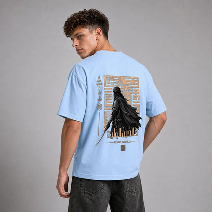 A man wearing a light blue t-shirt with a large back graphic featuring a black cloaked figure and gold geometric patterns, shown from the back against a gray studio background.