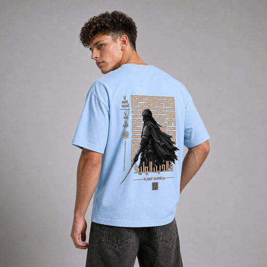 A man wearing a light blue t-shirt with a large back graphic featuring a black cloaked figure and gold geometric patterns, shown from the back against a gray studio background.