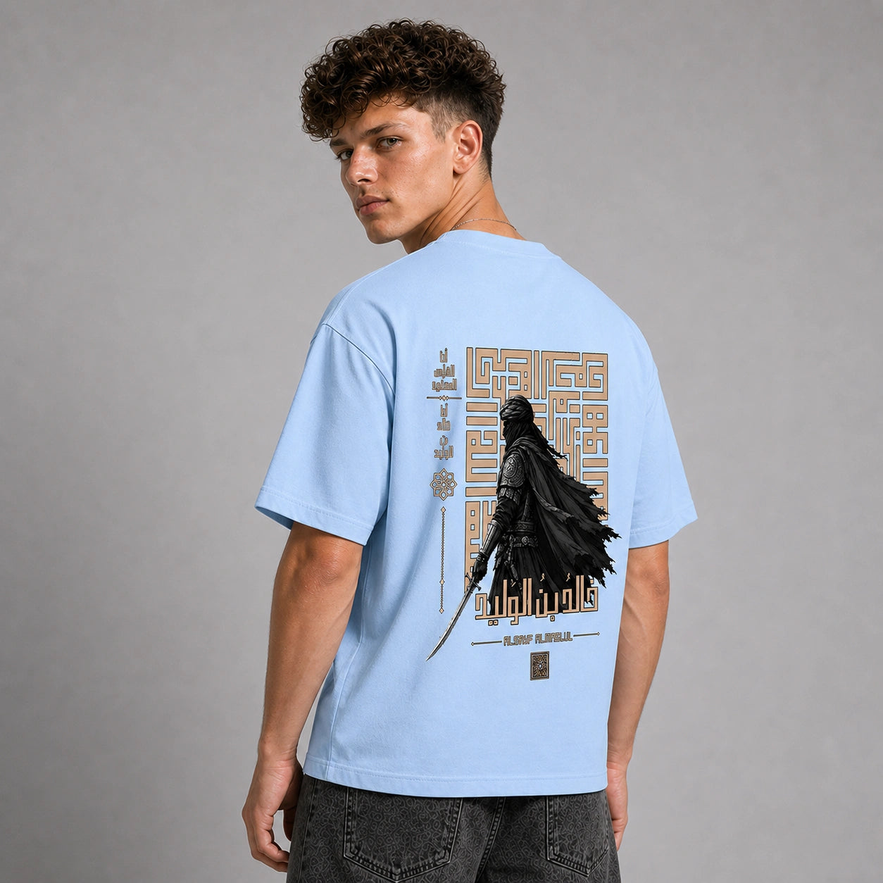 A man wearing a light blue t-shirt with a large graphic print on the back, featuring a black cloaked figure and gold geometric patterns, posed against a neutral gray studio background.