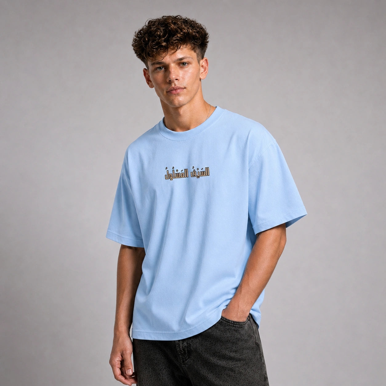 A young man with curly brown hair wearing a light blue crewneck t-shirt with a small graphic print on the chest, paired with dark gray jeans, standing against a neutral gray studio background.