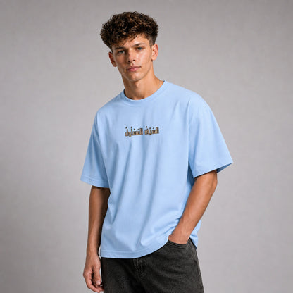 A young man with curly brown hair wearing a light blue crewneck t-shirt with a small graphic print on the chest, paired with dark gray jeans, standing against a neutral gray studio background.