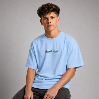 A young man with curly brown hair wearing a light blue t-shirt with a small graphic on the chest, sitting against a neutral gray studio background.