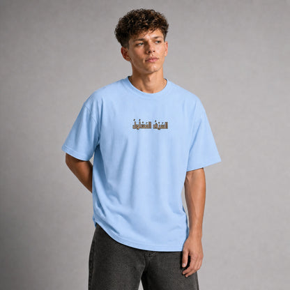 A man wearing a light blue crewneck t-shirt with a small graphic print on the chest, paired with dark gray jeans, standing against a neutral gray studio background.