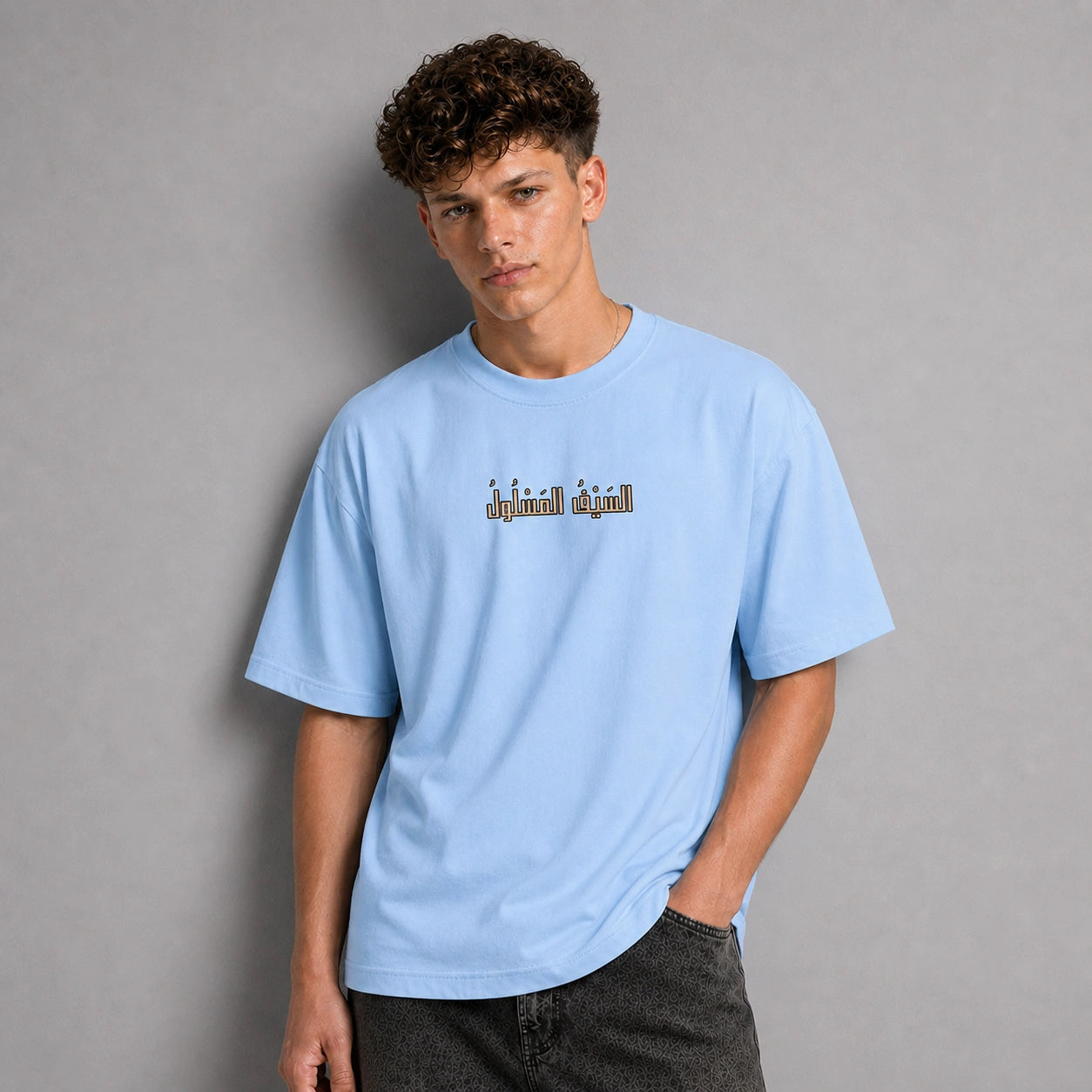 A young man with curly brown hair wearing a light blue t-shirt with a graphic text print on the chest, paired with dark gray jeans, standing against a solid gray studio background.