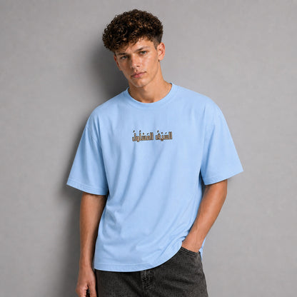 A young man with curly brown hair wearing a light blue t-shirt with a graphic text print on the chest, paired with dark gray jeans, standing against a solid gray studio background.