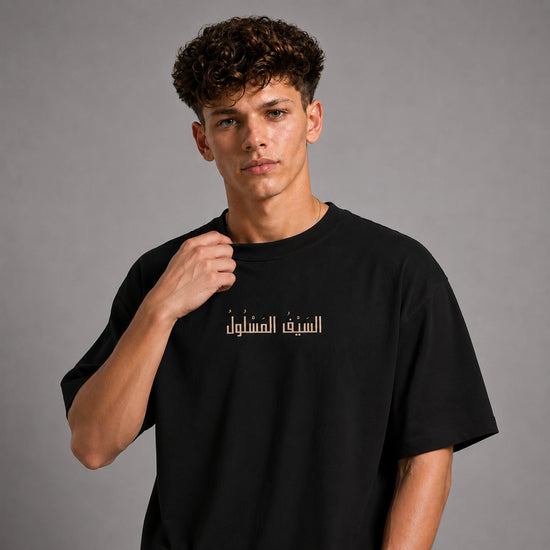 A young man with curly brown hair wearing a black short-sleeve t-shirt with gold Arabic calligraphy on the chest, posed against a neutral gray studio background.