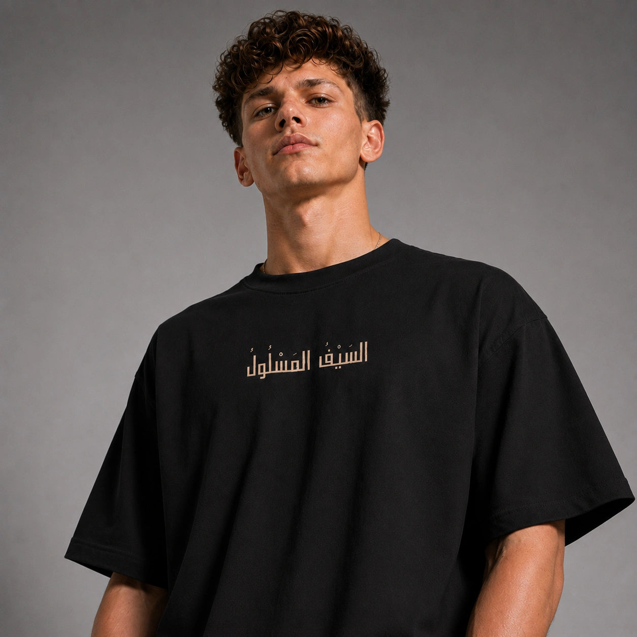 A man wearing a black oversized t-shirt with gold Arabic text printed across the chest, photographed against a neutral gray studio background.