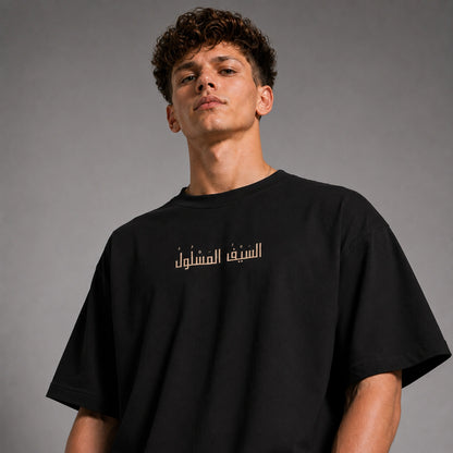 A man wearing a black oversized t-shirt with gold Arabic text printed across the chest, photographed against a neutral gray studio background.