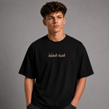 A young man with curly brown hair wearing a black crewneck t-shirt with Arabic text printed in beige across the chest, posed against a solid gray studio background.