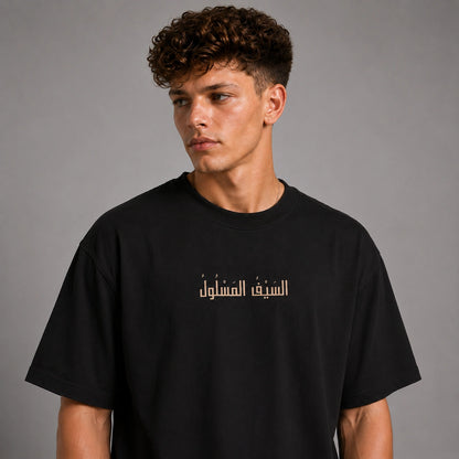 A young man with curly brown hair wearing a black crewneck t-shirt with gold Arabic text printed across the chest, photographed against a neutral gray studio background.