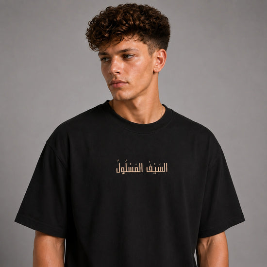 A young man with curly brown hair wearing a black crewneck t-shirt with gold Arabic text printed across the chest, photographed against a neutral gray studio background.