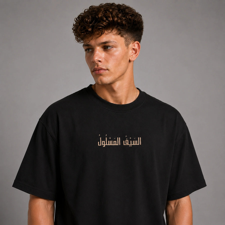 A young man with curly brown hair wearing a black crewneck t-shirt with gold Arabic text printed across the chest, photographed against a neutral gray studio background.
