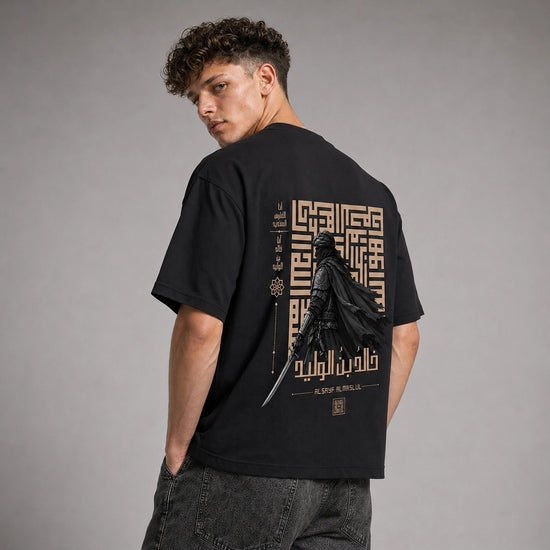 A man wearing a black t-shirt with a large back graphic featuring Darth Vader, Arabic calligraphy, and a gold geometric pattern, photographed against a neutral gray studio background.
